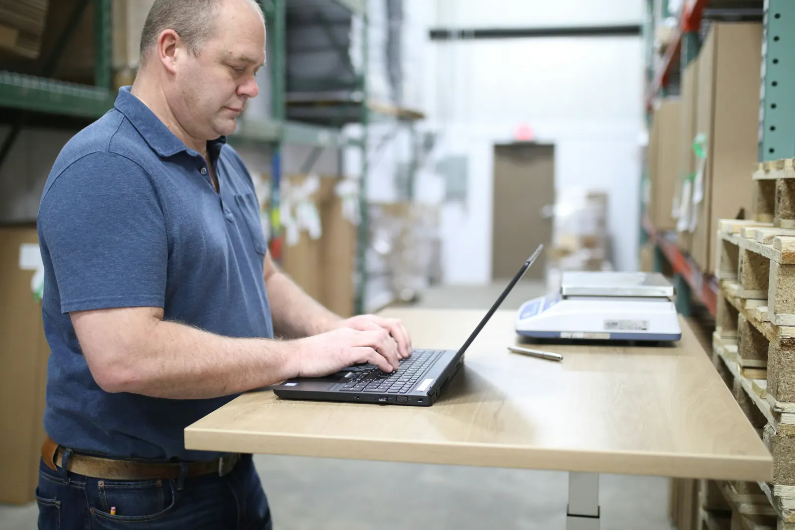 A dispatcher working at a laptop on a standing desk between warehouse racks.