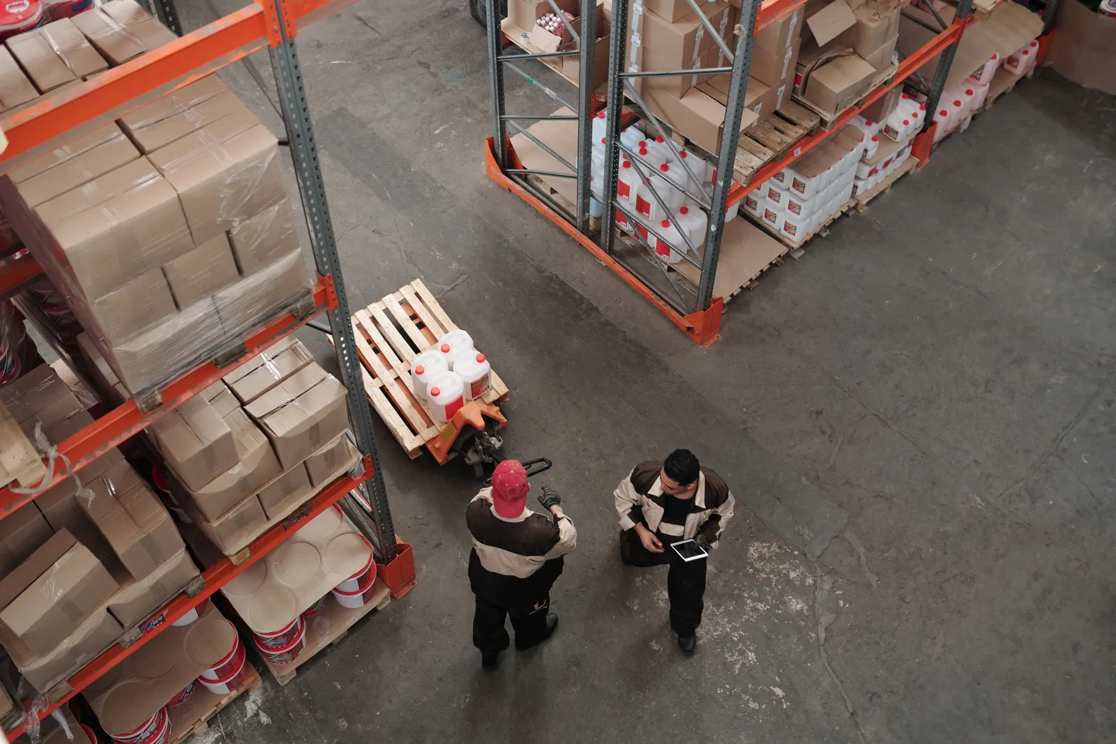 Two warehouse workers on the floor between racks, one holding a tablet, seen from above.